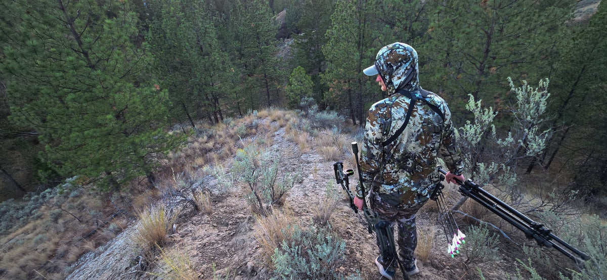 Bowhunter in Sitka camo carrying a compound bow and tripod along a pine ridge