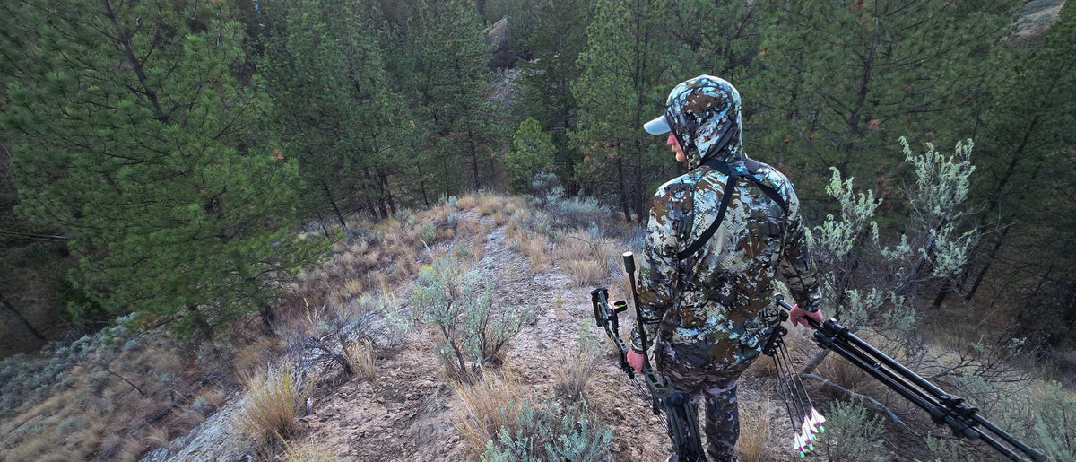 Bowhunter in Sitka camo carrying a compound bow and tripod along a pine ridge